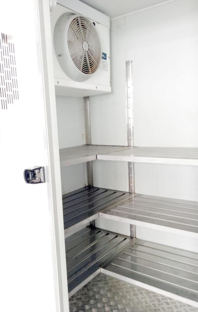 Intérieur Chambre Froide : Étagères et Évaporateur Intérieur d'une chambre froide ou d'un camion frigorifique, vue ouverte, montrant des étagères métalliques et un évaporateur avec ventilateur blanc en haut.