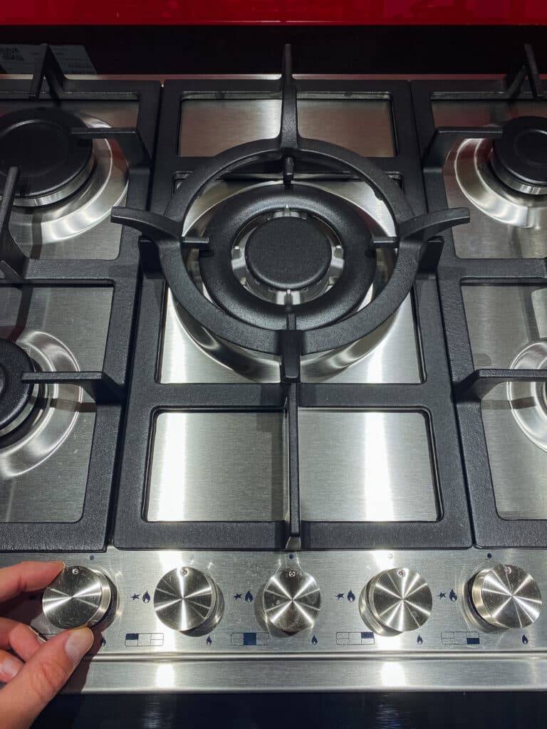 Main ajustant le bouton d'un fourneau à gaz inox à cinq brûleurs. Le grand brûleur central est visible au-dessus des commandes.