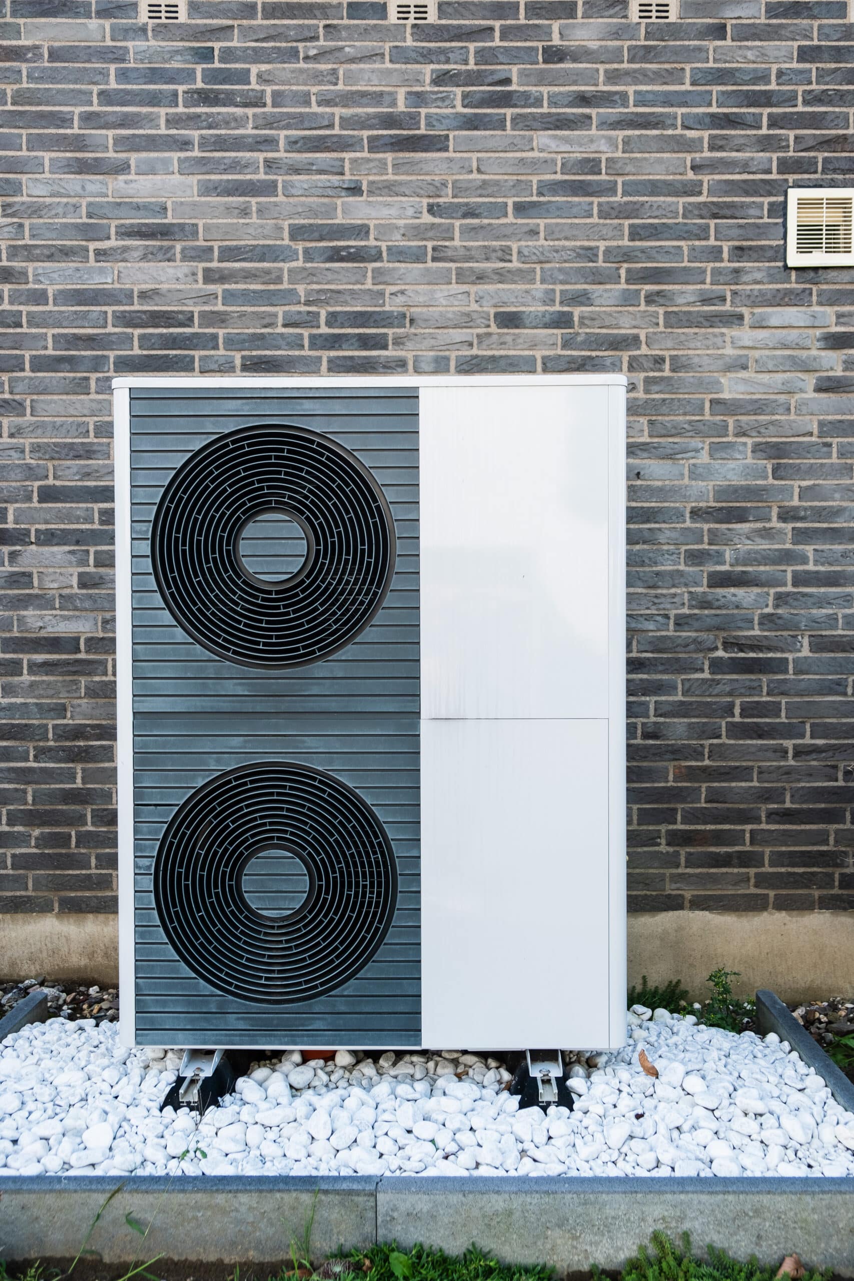 Pompe à chaleur extérieure : installation moderne sur galets blancs Unité extérieure de pompe à chaleur moderne, blanche et grise, installée devant un mur de briques grises texturées, reposant sur des galets blancs.
